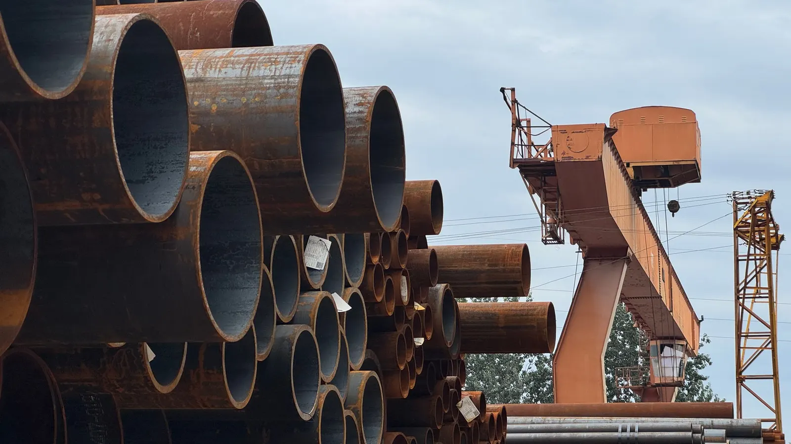 Stacked steel pipes of different diameters in an industrial yard beside a gantry crane