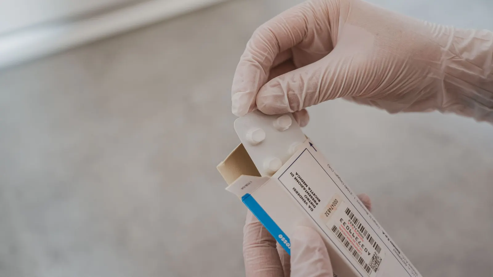 Gloved hands opening a prescription medicine carton with a barcode label and printed code, a blister pack sliding out