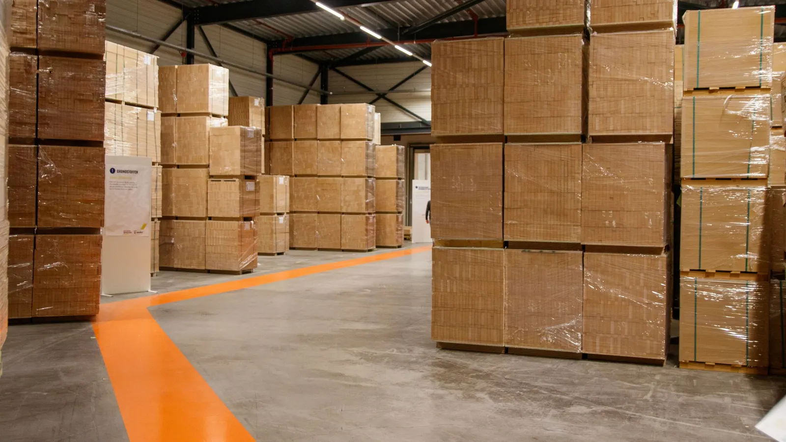 Warehouse floor with pallets of stacked cardboard boxes block-stacked either side of an orange marked walkway line