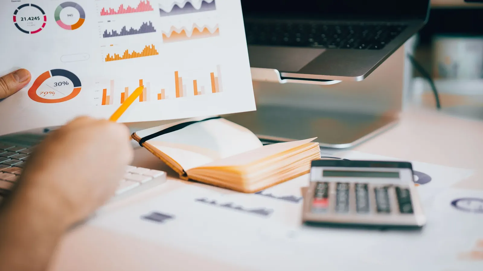 A hand holding a printed sheet of charts over a desk with a notebook, a calculator and a laptop