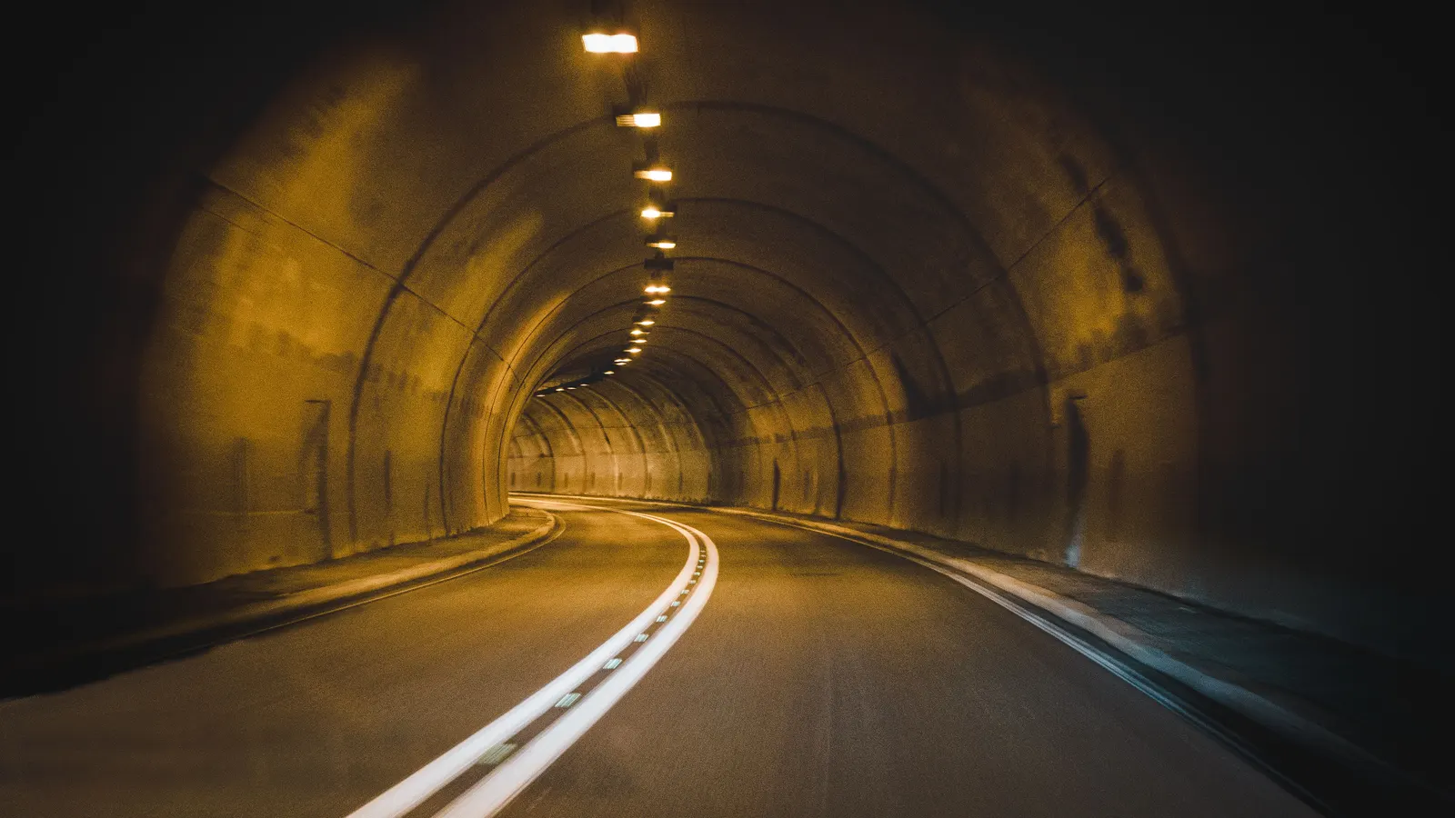 A curving road tunnel lit by a row of warm overhead lights, the only illumination between one signal and the next.