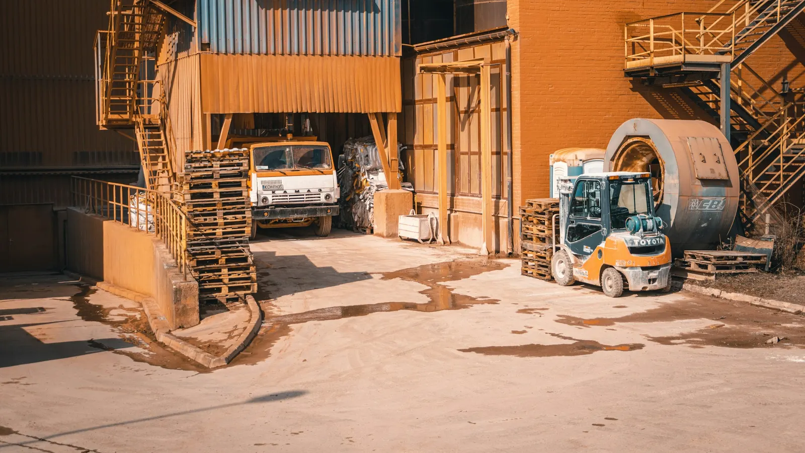 An old works truck parked under a loading structure in a factory yard, with stacked pallets and a forklift beside it