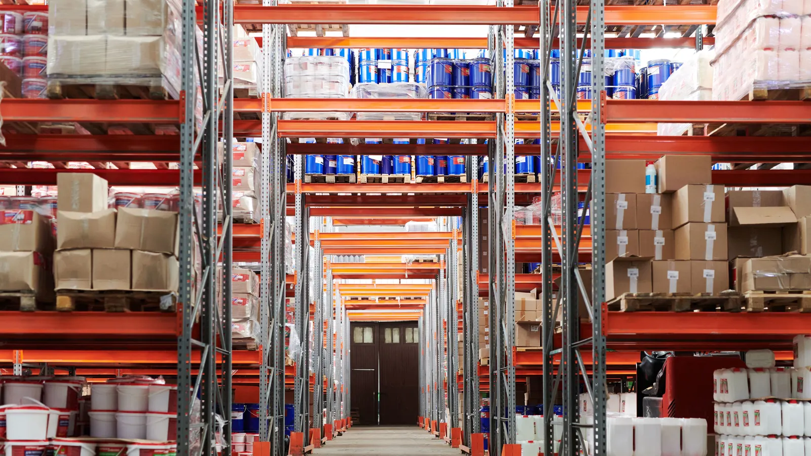 A long warehouse aisle between tall orange pallet racks stocked with boxes, drums and buckets