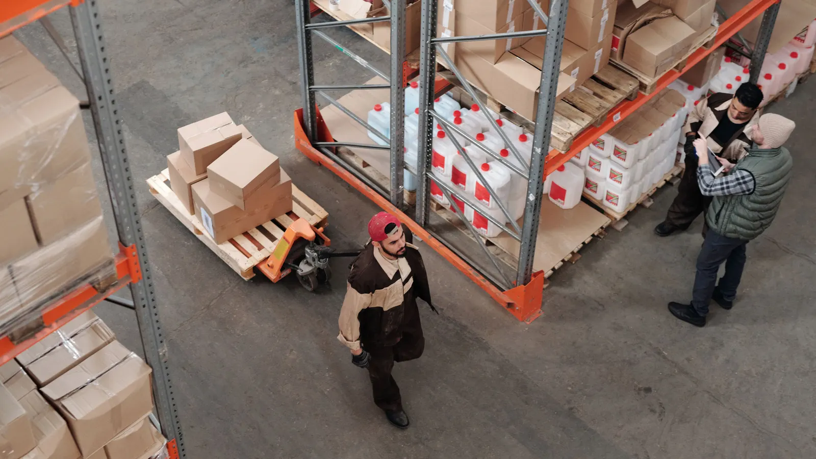 Corsia di magazzino vista dall’alto: un addetto traina un transpallet con scatoloni mentre due colleghi controllano le giacenze alla scaffalatura